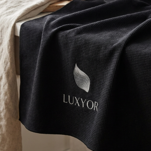 Zwarte Luxyor inlegdeken met geborduurd logo over het voeteneinde van een bed